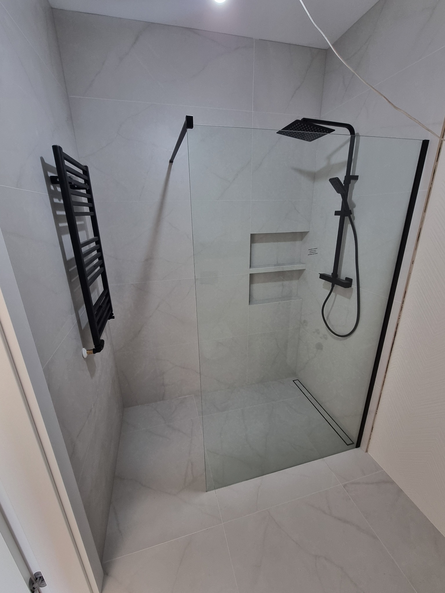 Nowoczesna kabina prysznicowa typu walk-in z czarnymi elementami wykończeniowymi, grzejnik drabinkowy w kolorze czarnym, szare płytki imitujące marmur, odpływ liniowy w podłodze, Warszawa.