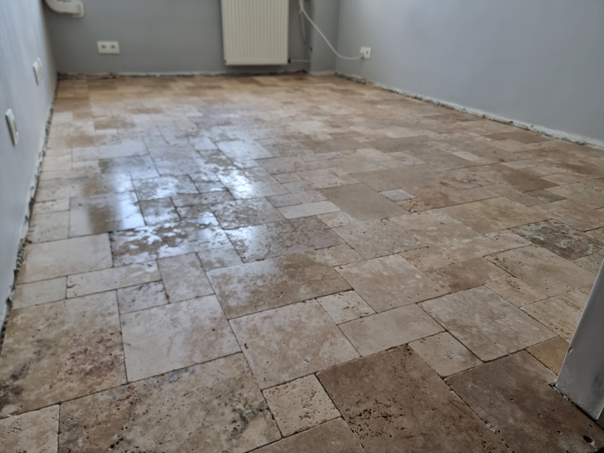 Nowo ułożona, beżowa podłoga z trawertynu w pomieszczeniu z szarymi ścianami i białym grzejnikiem, widoczne niedociągnięcia przy ścianach.