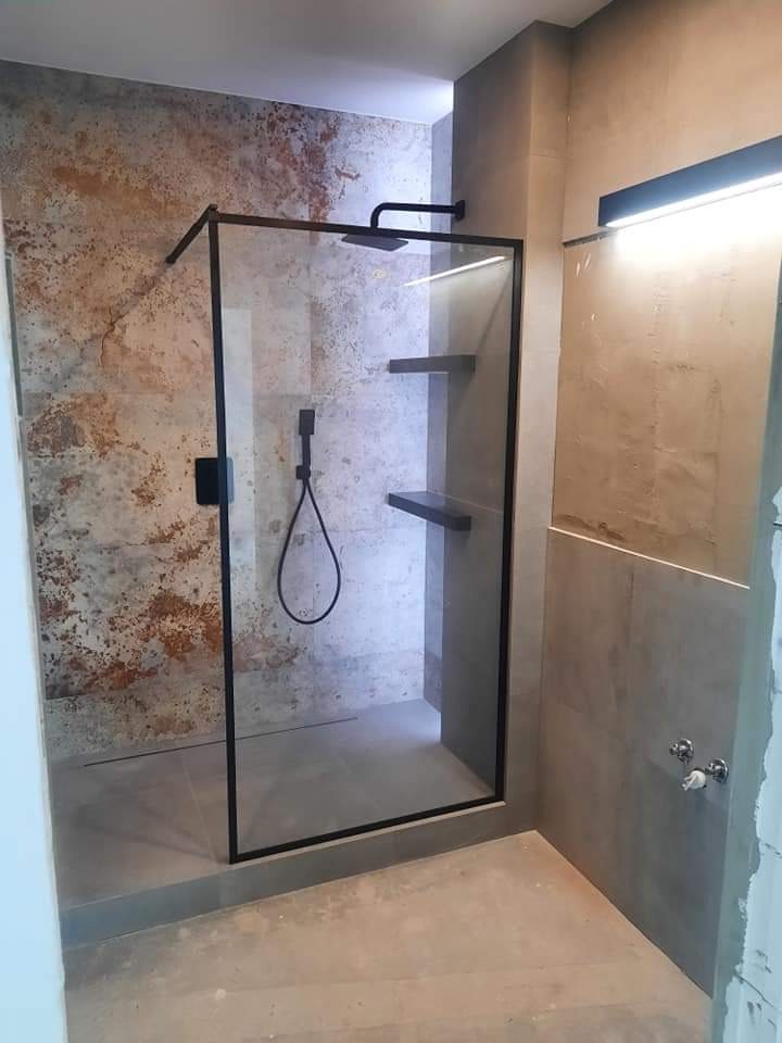 Nowoczesna kabina prysznicowa z czarną ramą i deszczownicą, półki wewnątrz, ściana wykończona płytkami imitującymi rdzę, szara podłoga i oświetlenie LED.