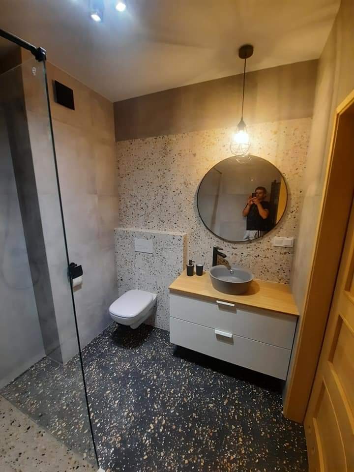 Nowoczesna łazienka z kabiną prysznicową, umywalką nablatową w kolorze szarym, szafką z drewnianym blatem i okrągłym lustrem z odbiciem fotografa, wykończona płytkami lastryko na ścianach i podłodze.