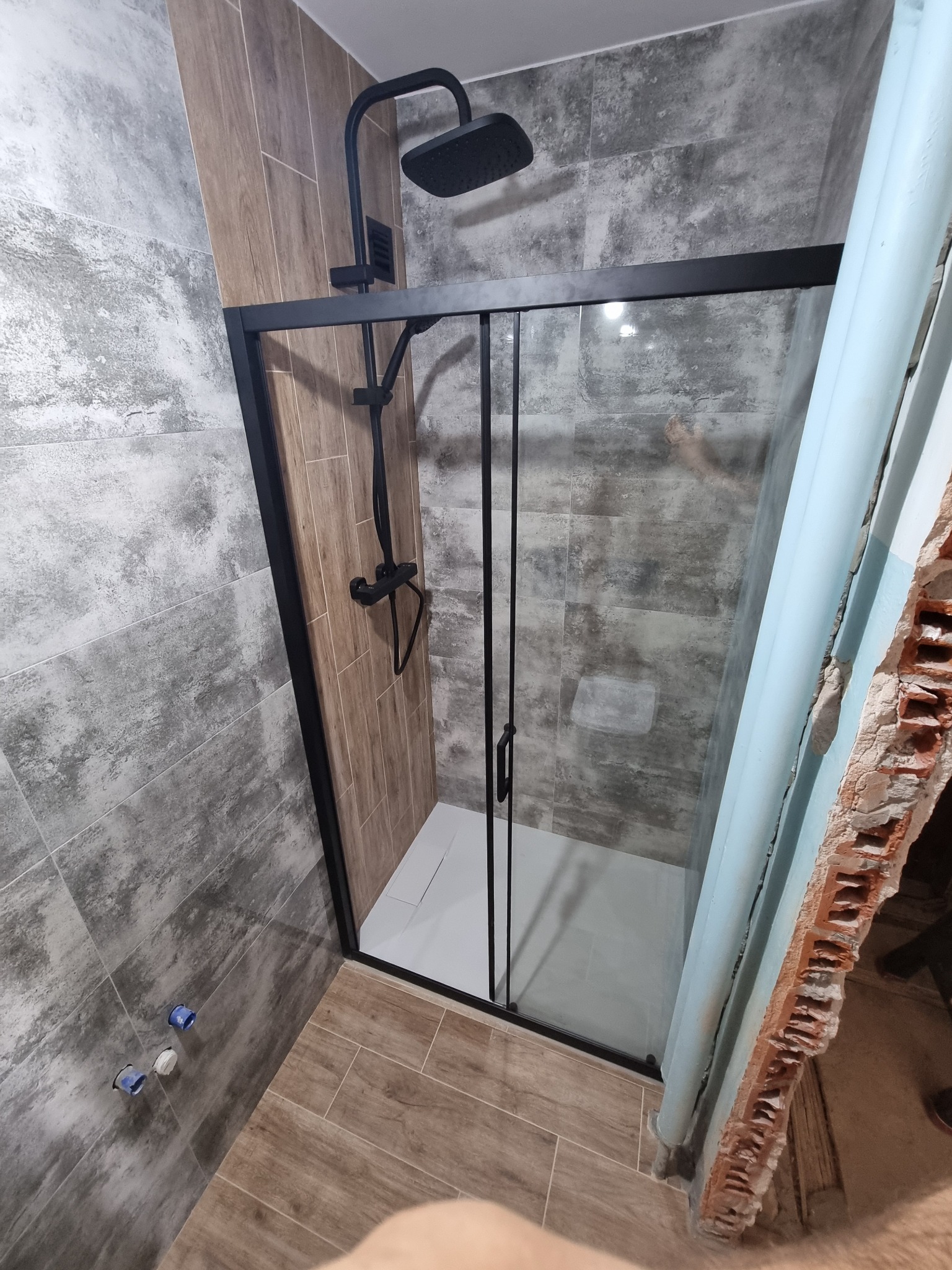 Kabina prysznicowa z czarną ramą i deszczownicą w łazience z szaro-brązowymi płytkami, widoczne niedokończone wykończenie ściany z cegły.
