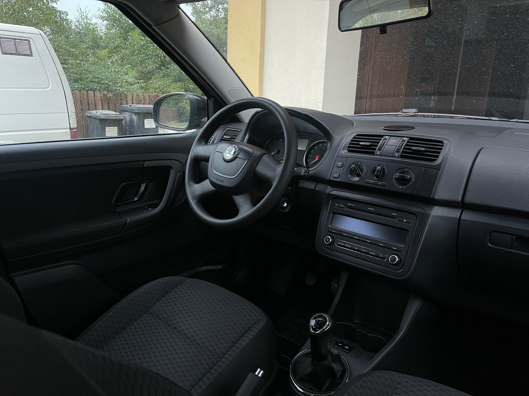 Wnętrze samochodu marki Skoda, widok na deskę rozdzielczą, kierownicę z logo, radio samochodowe i dźwignię zmiany biegów; fotel kierowcy z ciemną tapicerką.