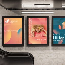 Arabeska Art & Brand Design - Trzy plakaty reklamowe Centrum Hildegardy Wrocław umieszczone na ścianie w pobliżu schodów ruchomych. Plakaty w kolorach pomarańczowym, różowym i niebieskim, z różnymi hasłami i grafikami.