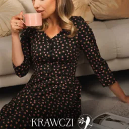 Kobieta pije z różowego kubka, ubrana w sukienkę w drobne kwiatki, obok leży otwarty katalog marki odzieżowej KRAWCZI z logo przedstawiającym ptaka na gałęzi.
