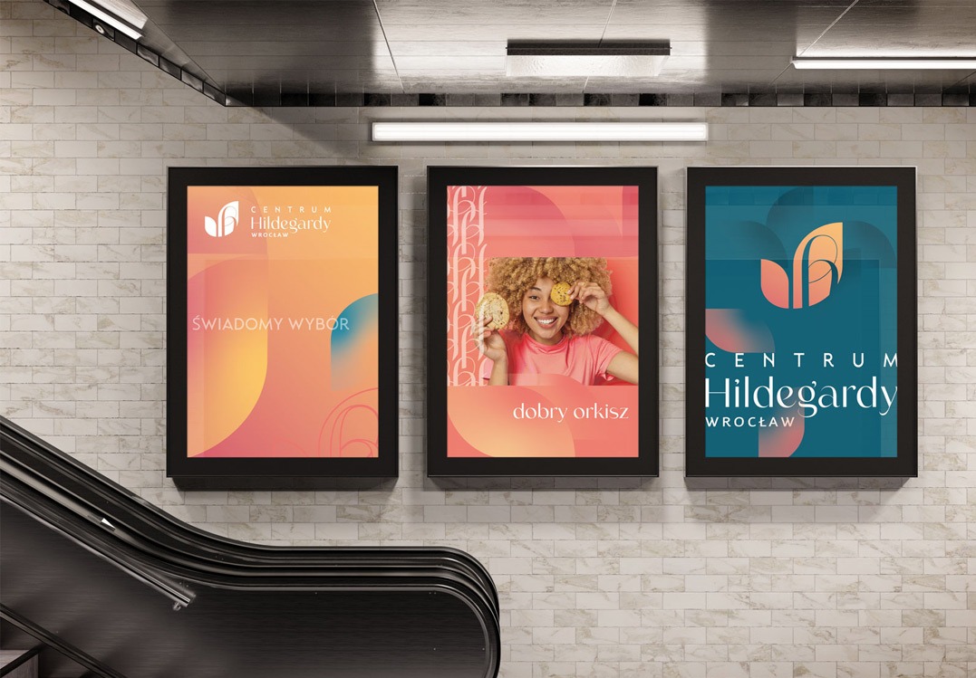 Trzy plakaty reklamowe Centrum Hildegardy Wrocław umieszczone na ścianie w pobliżu schodów ruchomych. Plakaty w kolorach pomarańczowym, różowym i niebieskim, z różnymi hasłami i grafikami.