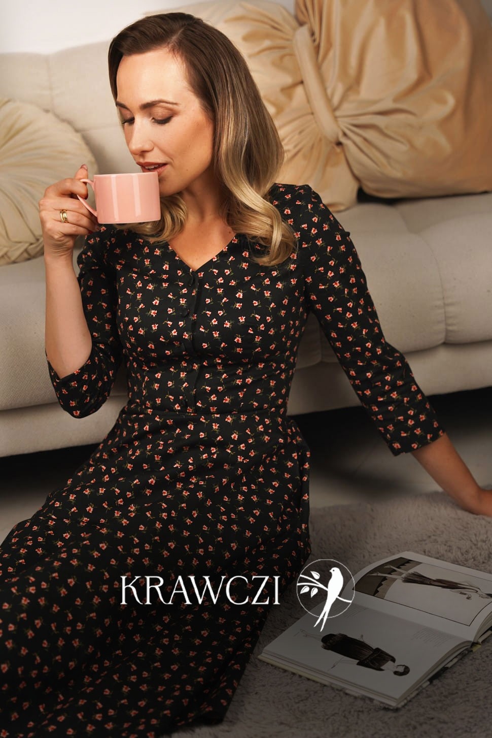 Kobieta pije z różowego kubka, ubrana w sukienkę w drobne kwiatki, obok leży otwarty katalog marki odzieżowej KRAWCZI z logo przedstawiającym ptaka na gałęzi.