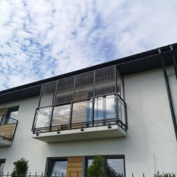 Grzegorz Ziomek Kotiery - Balkon z siatką zabezpieczającą dla kota, widok z dołu na jasny budynek z balkonami, siatka zamontowana bezinwazyjnie na ramie z czarnego metalu, w tle błękitne niebo z chmurami.