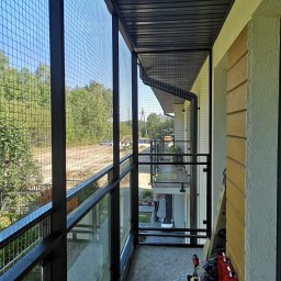 Grzegorz Ziomek Kotiery - Zabezpieczenie balkonu siatką stalową, widok z wewnątrz balkonu na zewnątrz z widocznym lasem w tle, czarne metalowe słupki i szyny, częściowo widoczne narzędzia i akcesoria montażowe.