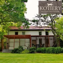 Przydomowa konstrukcja drewniana, prawdopodobnie woliera dla kotów, przylegająca do białego domu z czerwonym dachem, otoczona zielenią i drzewami. W prawym górnym rogu widoczne logo firmy.