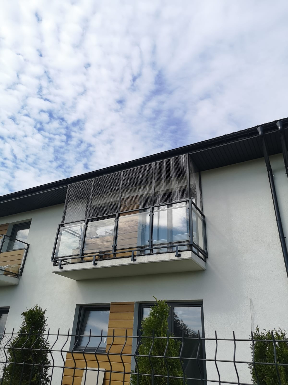 Balkon z siatką zabezpieczającą dla kota, widok z dołu na jasny budynek z balkonami, siatka zamontowana bezinwazyjnie na ramie z czarnego metalu, w tle błękitne niebo z chmurami.