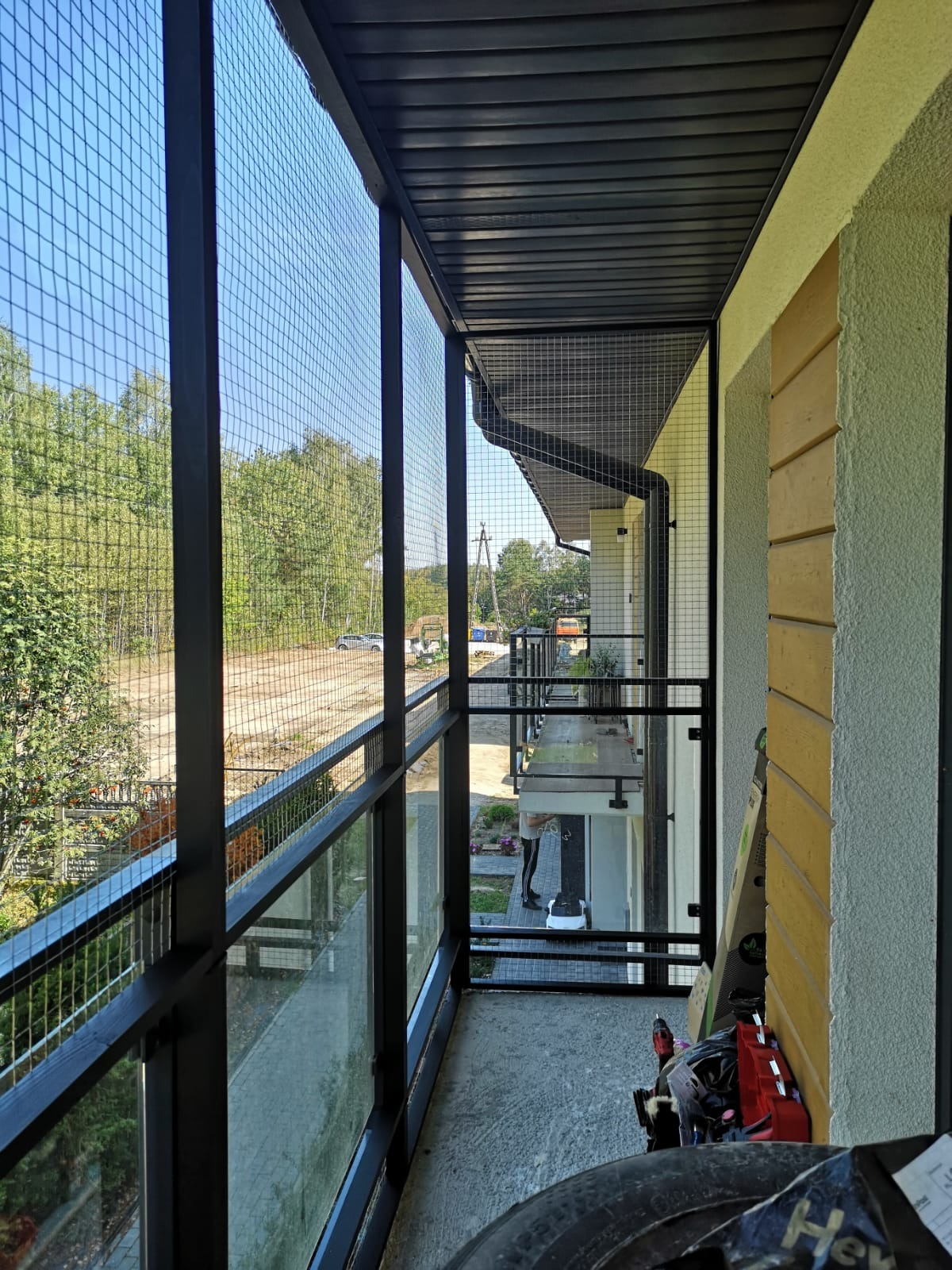 Zabezpieczenie balkonu siatką stalową, widok z wewnątrz balkonu na zewnątrz z widocznym lasem w tle, czarne metalowe słupki i szyny, częściowo widoczne narzędzia i akcesoria montażowe.