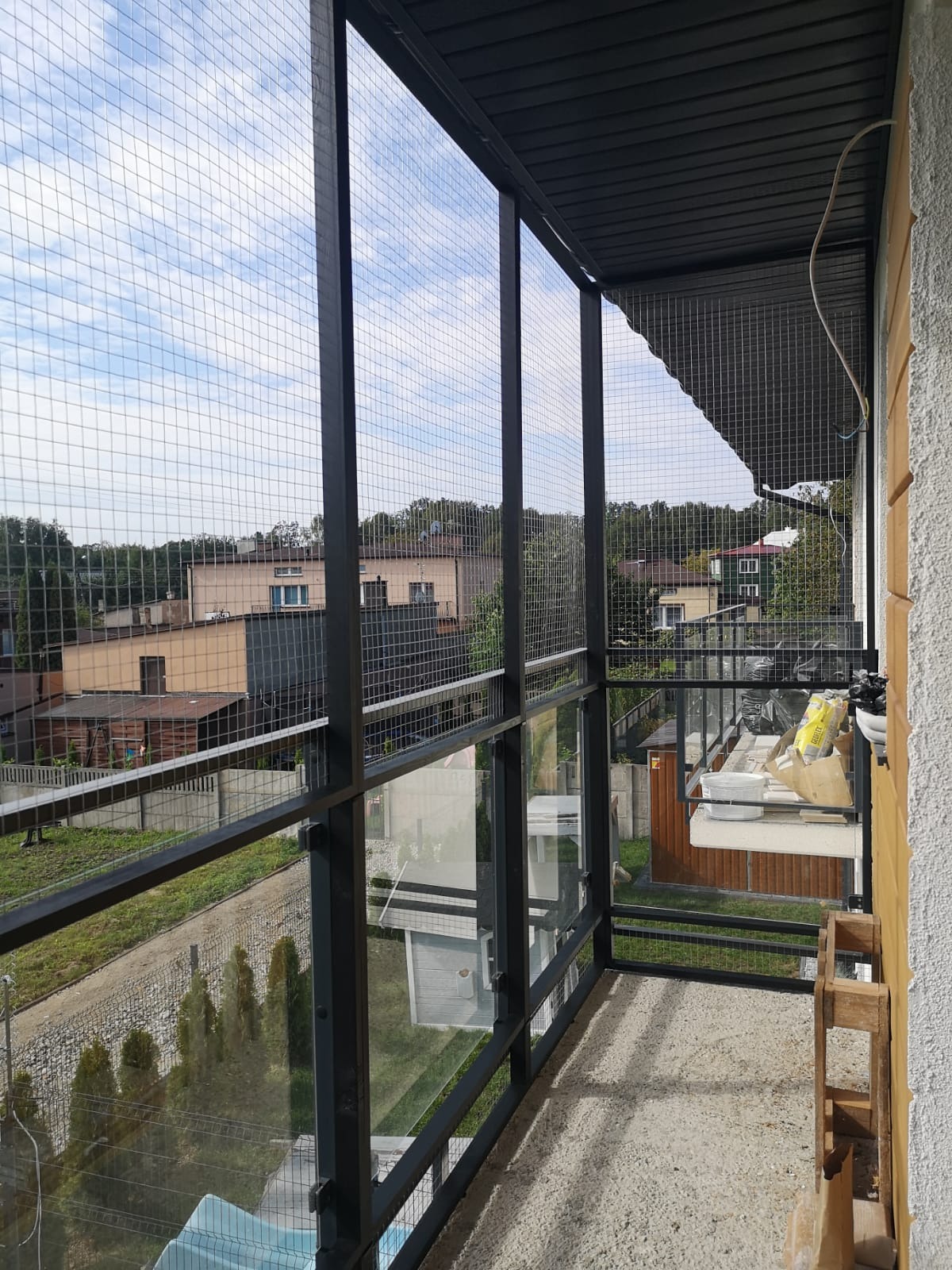 Zabezpieczenie balkonu siatką stalową w czarnej ramie, widok z góry na okoliczne budynki i zieleń.