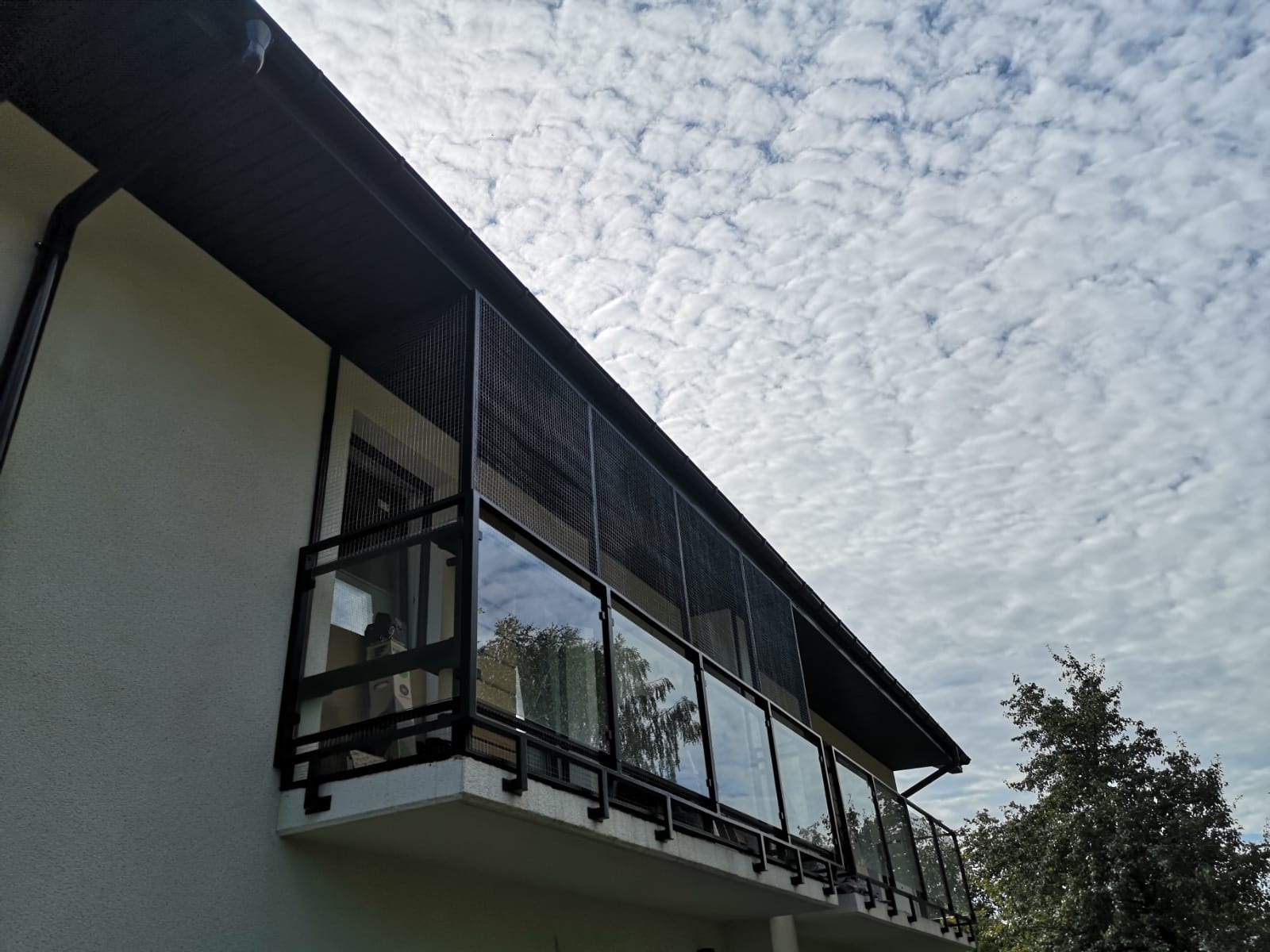 Zabezpieczenie balkonu siatką stalową, widok z dołu na balkon z czarną ramą i szklanymi panelami, siatka ochronna dla zwierząt zamontowana na całej długości balkonu, na tle pochmurnego nieba.