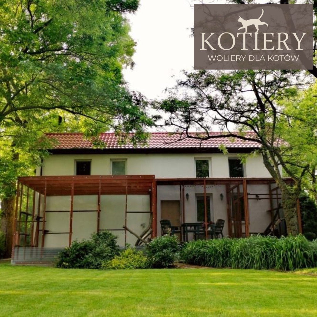 Przydomowa konstrukcja drewniana, prawdopodobnie woliera dla kotów, przylegająca do białego domu z czerwonym dachem, otoczona zielenią i drzewami. W prawym górnym rogu widoczne logo firmy.