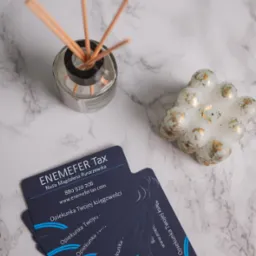 Wizytówki biura księgowego ENEMEFER Tax z Poznania, ułożone wachlarzowo na marmurowym tle, obok świeczki w kształcie kuli i dyfuzora zapachowego z patyczkami.