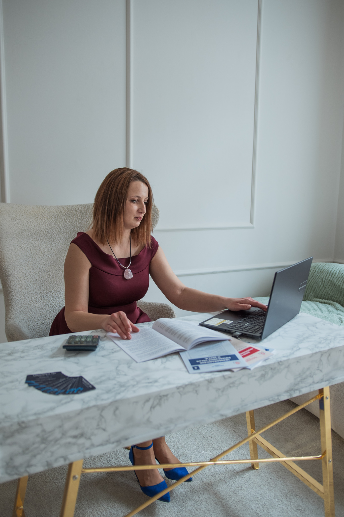 Kobieta w bordowej sukience pracuje przy biurku z laptopem, kalkulatorem i otwartą książką, na biurku leżą wizytówki ułożone wachlarzowo, a na nogach ma niebieskie buty na obcasie.