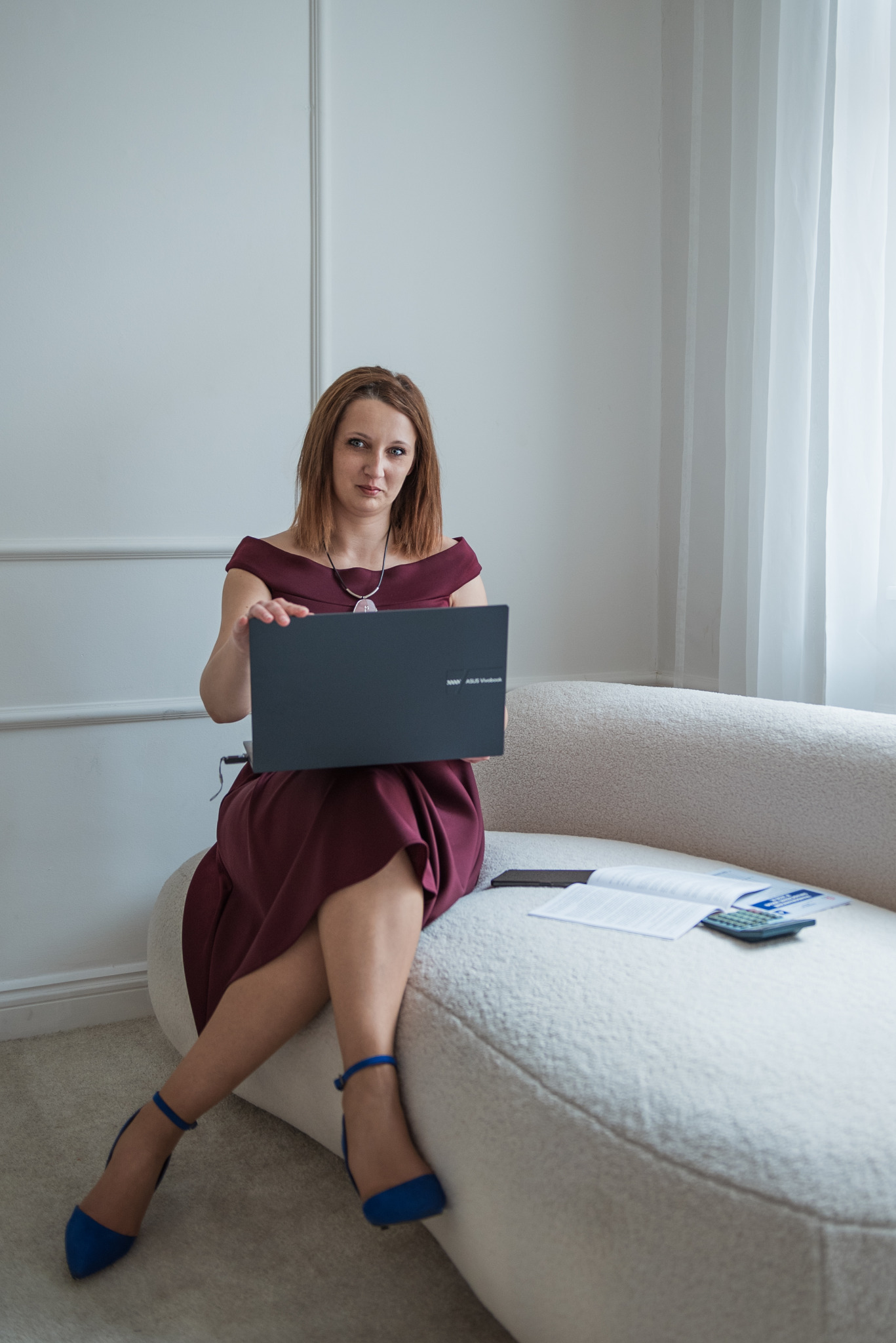 Kobieta w bordowej sukience i niebieskich szpilkach siedzi na białej sofie, trzymając otwarty laptop Asus Vivobook; obok kalkulator i dokumenty.