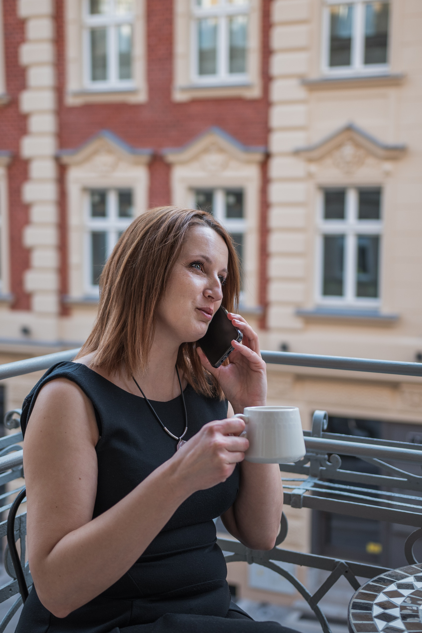 Kobieta w czarnej sukience rozmawia przez telefon na balkonie, trzymając biały kubek. W tle kamienica z białymi oknami.
