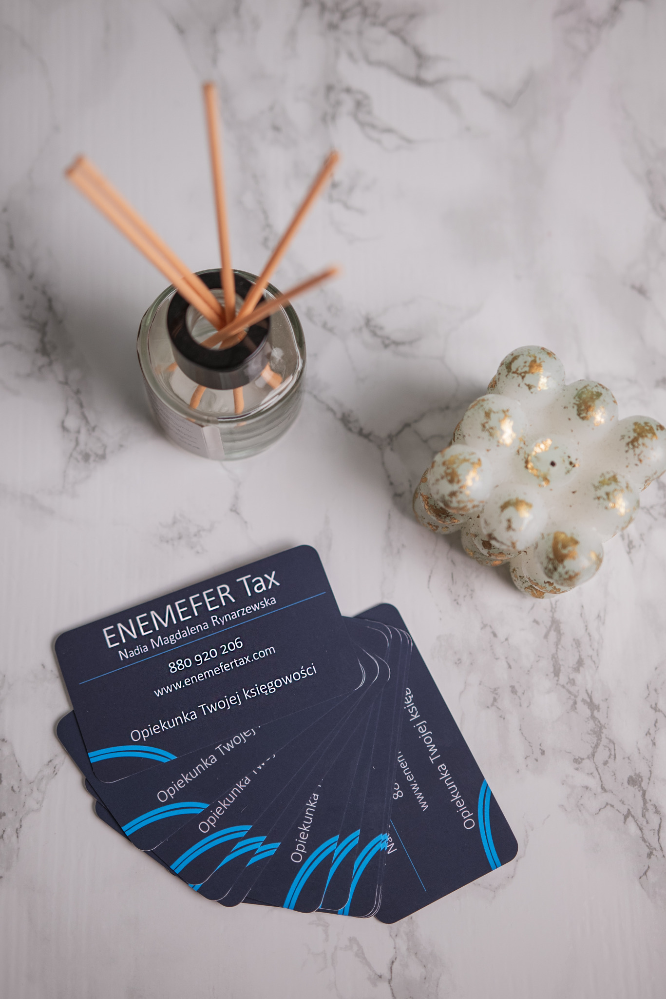 Wizytówki biura księgowego ENEMEFER Tax z Poznania, ułożone wachlarzowo na marmurowym tle, obok świeczki w kształcie kuli i dyfuzora zapachowego z patyczkami.