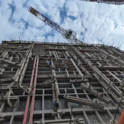 Szare szalunki z betonu zbrojonego na budowie, widok z dołu na konstrukcję i żuraw budowlany na tle pochmurnego nieba.