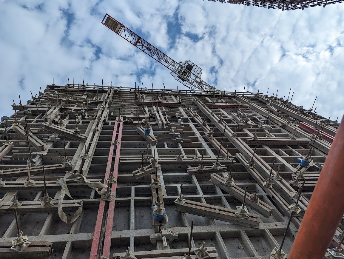 Szare szalunki z betonu zbrojonego na budowie, widok z dołu na konstrukcję i żuraw budowlany na tle pochmurnego nieba.