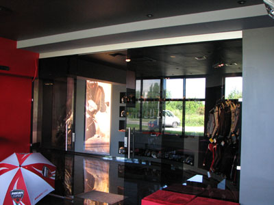 Szklane drzwi przesuwne w salonie motocyklowym Ducati, z widocznymi elementami ekspozycji motocyklowej odzieży i akcesoriów, odbijające światło w polerowanej podłodze.