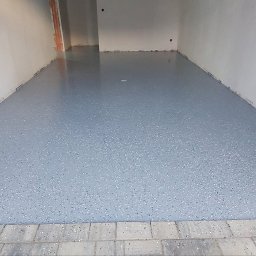 Mikrocement floor Design - Jasnoszara, gładka posadzka z drobnymi, ciemnymi i białymi płatkami dekoracyjnymi, wykończona na wysoki połysk, widoczna w surowym wnętrzu z białymi ścianami i odkrytymi instalacjami elektrycznymi.