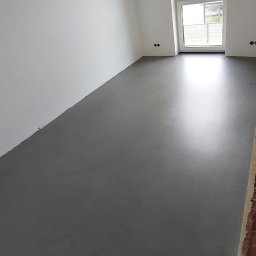 Mikrocement floor Design - Szara, jednolita podłoga w jasnym pomieszczeniu z białymi ścianami i oknem z widokiem na zewnątrz.