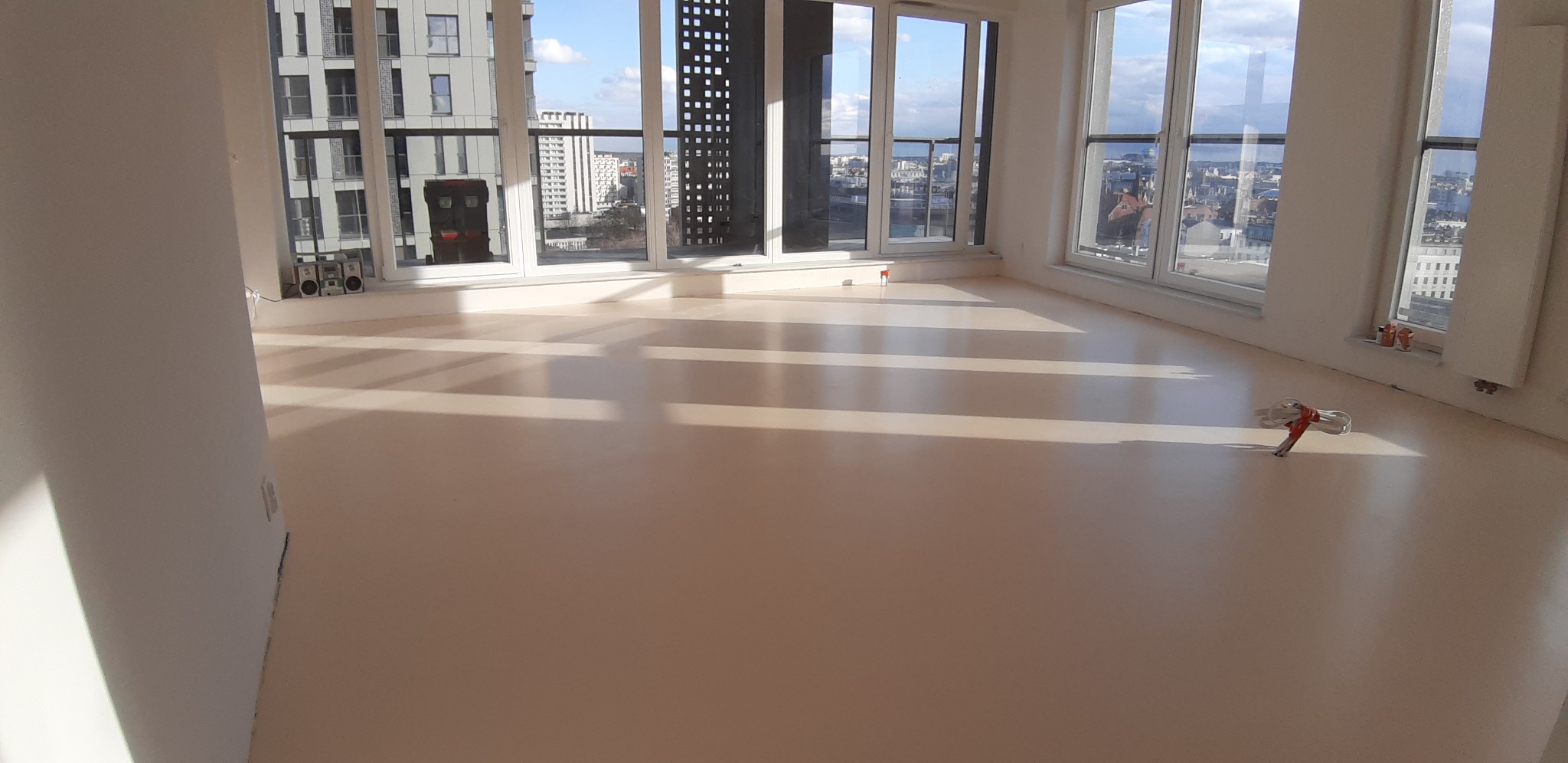 Jasne, jednolite wykończenie podłogi w nowym apartamencie z dużymi oknami i widokiem na miasto; na podłodze leży zwinięty kabel.