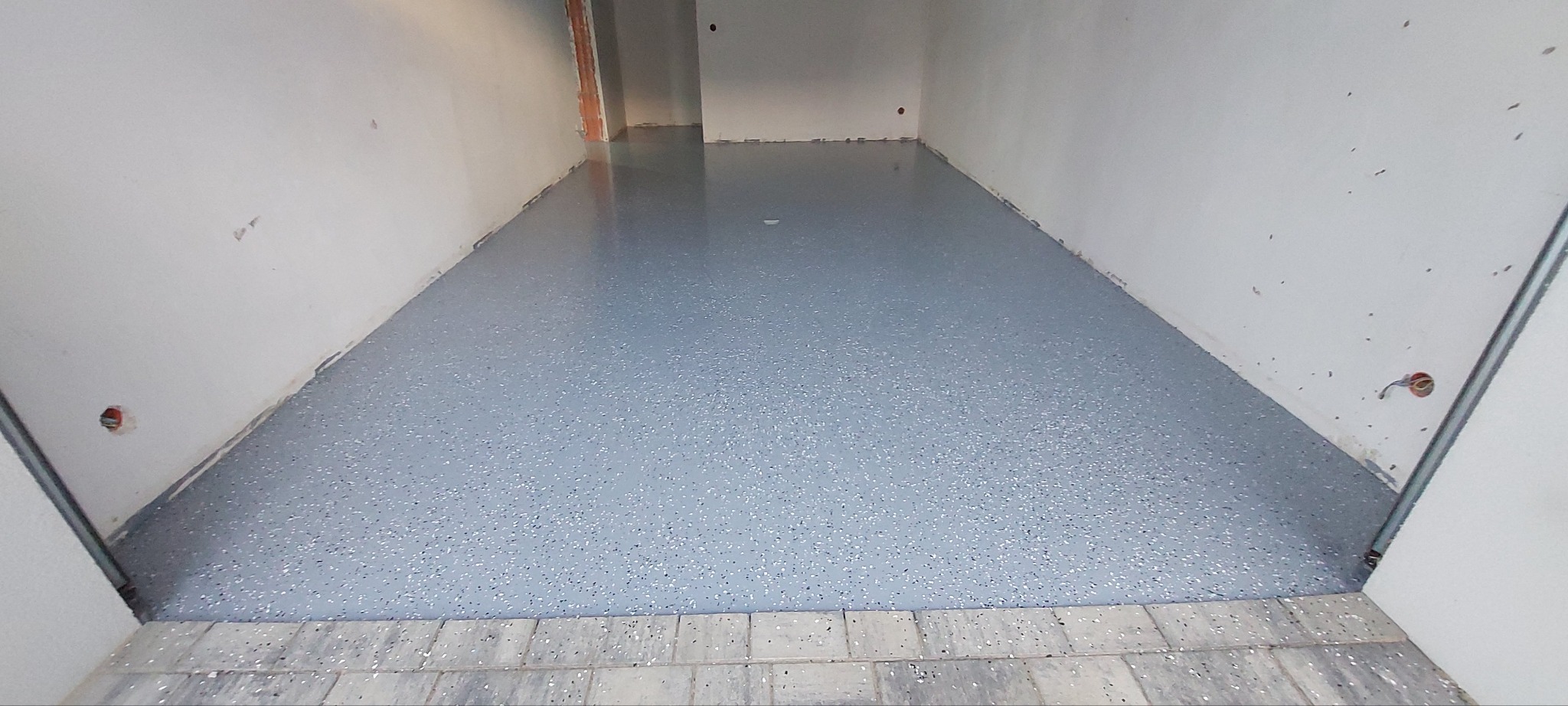 Jasnoszara, gładka posadzka z drobnymi, ciemnymi i białymi płatkami dekoracyjnymi, wykończona na wysoki połysk, widoczna w surowym wnętrzu z białymi ścianami i odkrytymi instalacjami elektrycznymi.