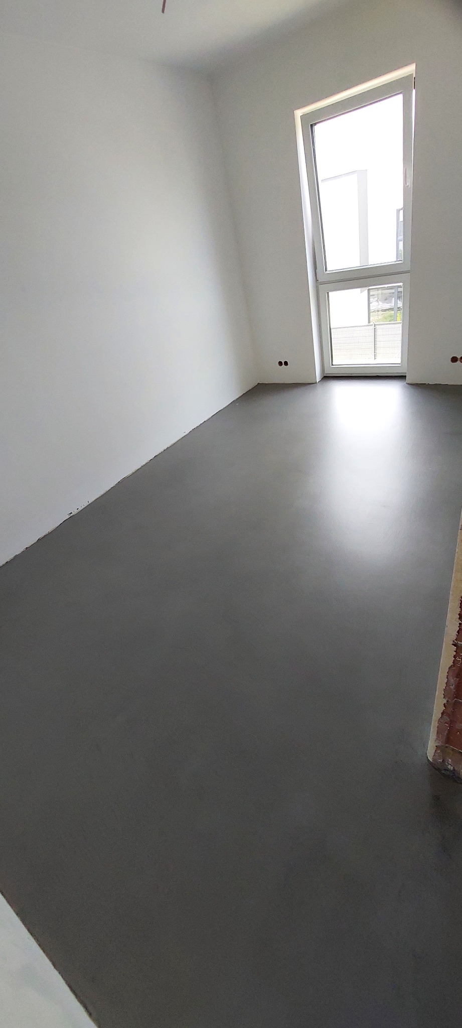 Szara, jednolita podłoga w jasnym pomieszczeniu z białymi ścianami i oknem z widokiem na zewnątrz.