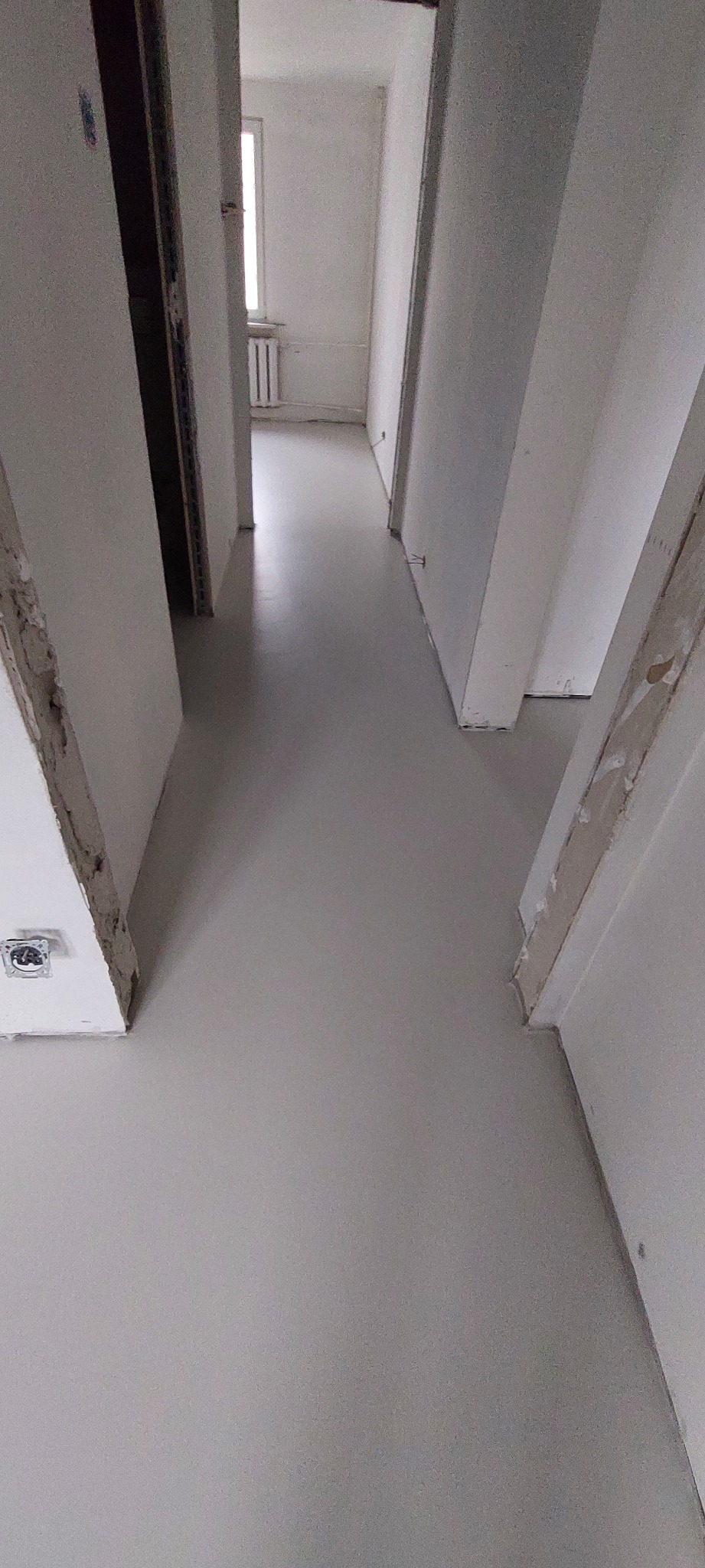 Szare, jednolite wykończenie podłogi w korytarzu mieszkania w trakcie remontu, widoczne niedokończone obramowania drzwi i gniazdko elektryczne bez osprzętu.
