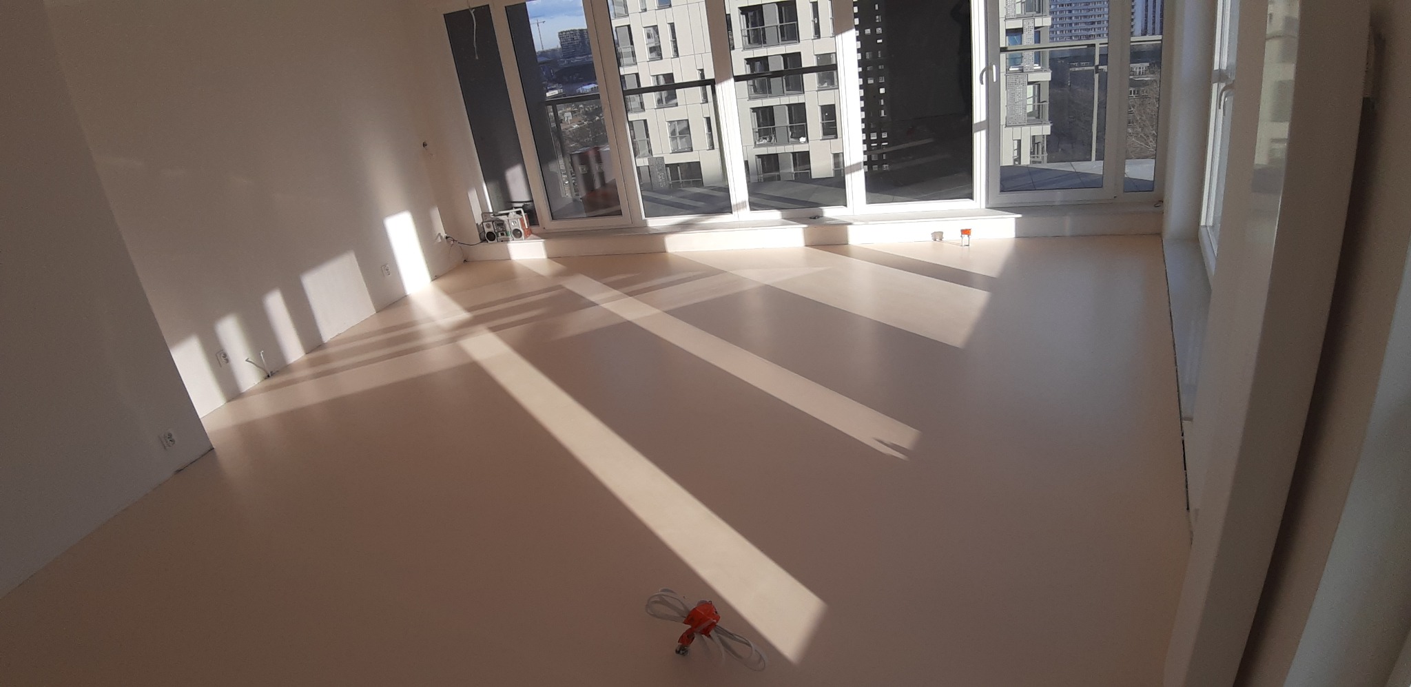 Jasna, wylewana posadzka w nowym mieszkaniu, z widocznymi promieniami słońca wpadającymi przez okna i budynkami w tle, mały boombox stoi przy ścianie, a na podłodze leżą zwinięte kable.