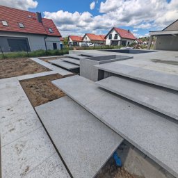 Sebbruk Sebastian Jurkiewicz - Szare płyty granitowe układane na tarasie domu jednorodzinnego w słoneczny dzień. Widoczne fragmenty podłoża i pobliskie budynki z czerwoną dachówką.