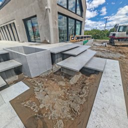 Sebbruk Sebastian Jurkiewicz - Montaż granitowych schodów i obrzeży basenu przy nowoczesnym domu. Widoczny mini-ciągnik i paleta z elementami. Prace w toku, teren przygotowany pod kostkę.