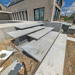 Sebbruk Sebastian Jurkiewicz - Granitowe schody zewnętrzne prowadzące do nowoczesnego domu z dużymi oknami. Widoczne płyty granitowe i elementy konstrukcyjne w trakcie montażu na zewnątrz budynku.