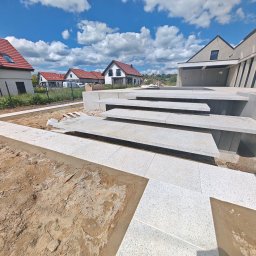 Sebbruk Sebastian Jurkiewicz - Nowoczesne schody z jasnej kostki granitowej prowadzą do domu na tle błękitnego nieba z chmurami. Widoczny fragment tarasu w trakcie budowy i domy w oddali.