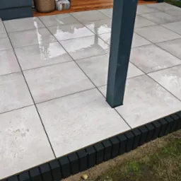Szare, kwadratowe płyty tarasowe ułożone na zewnątrz, obrzeża z ciemnych palików, szara kolumna. Widoczne ślady po deszczu.