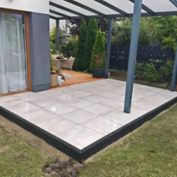 Nowoczesny taras z jasnych, kwadratowych płyt, obramowany czarnym, palisadowym obrzeżem. Zadaszenie pergoli, drewniany podest w tle i zielony trawnik.