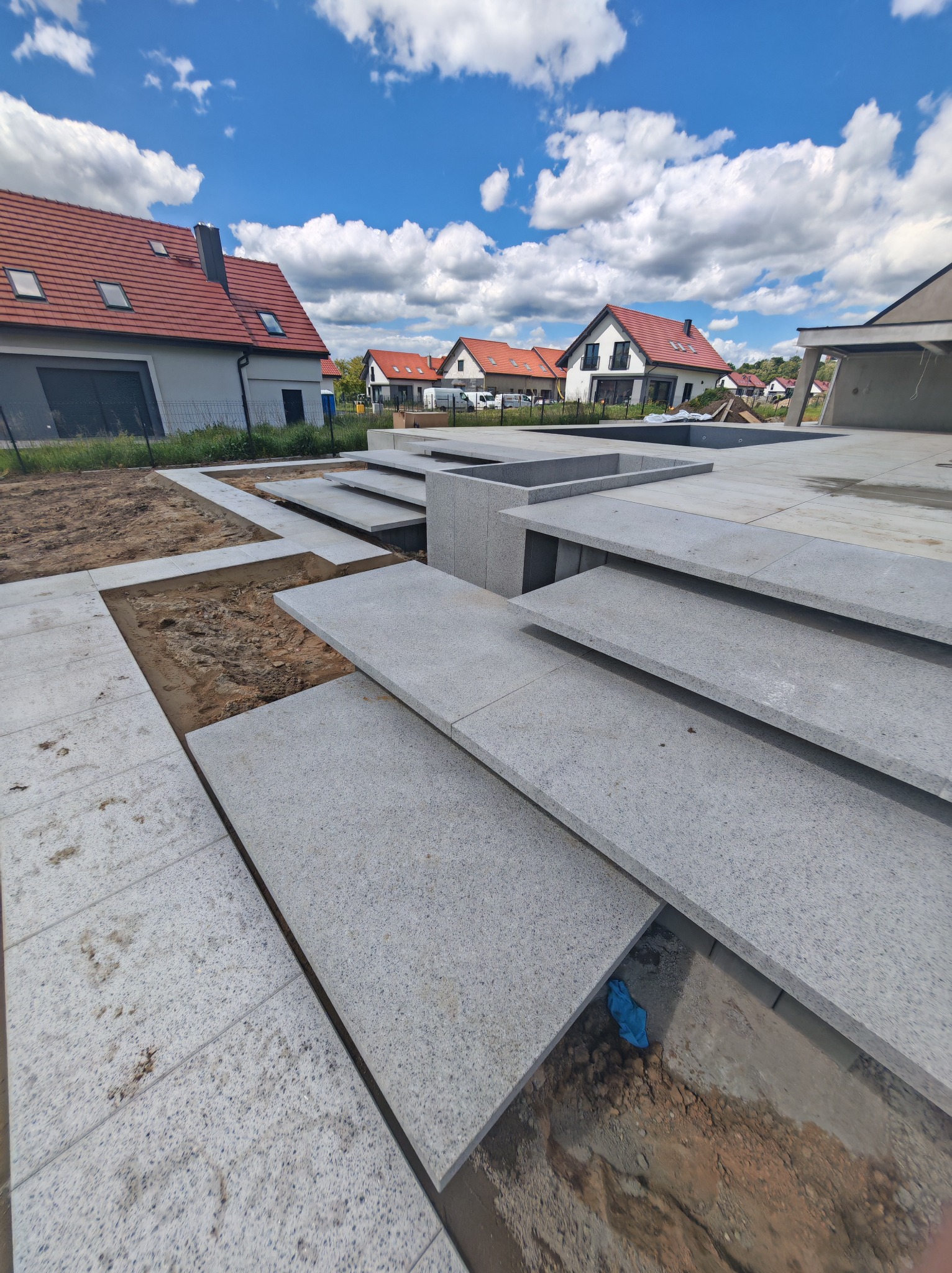 Szare płyty granitowe układane na tarasie domu jednorodzinnego w słoneczny dzień. Widoczne fragmenty podłoża i pobliskie budynki z czerwoną dachówką.