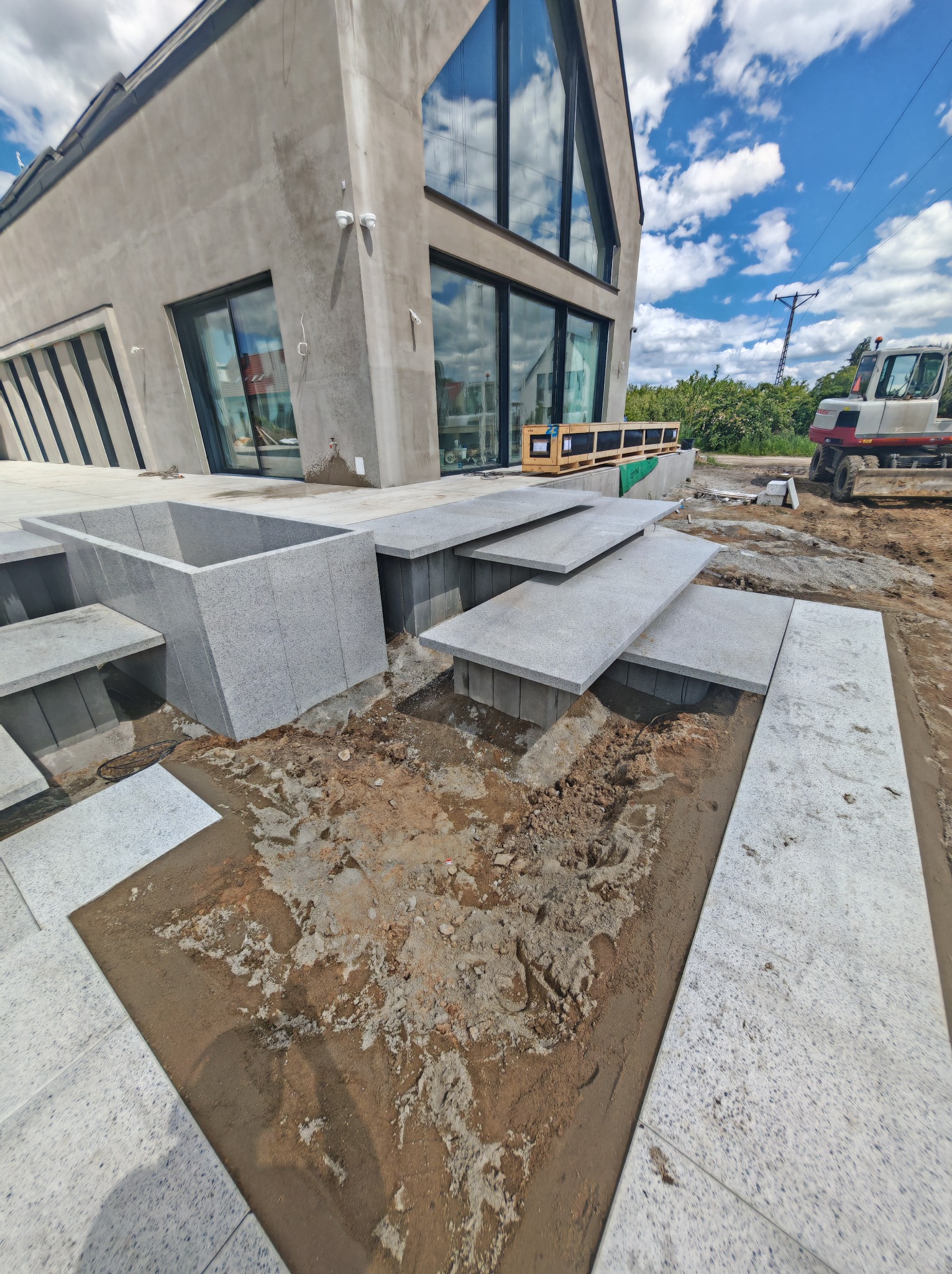 Montaż granitowych schodów i obrzeży basenu przy nowoczesnym domu. Widoczny mini-ciągnik i paleta z elementami. Prace w toku, teren przygotowany pod kostkę.