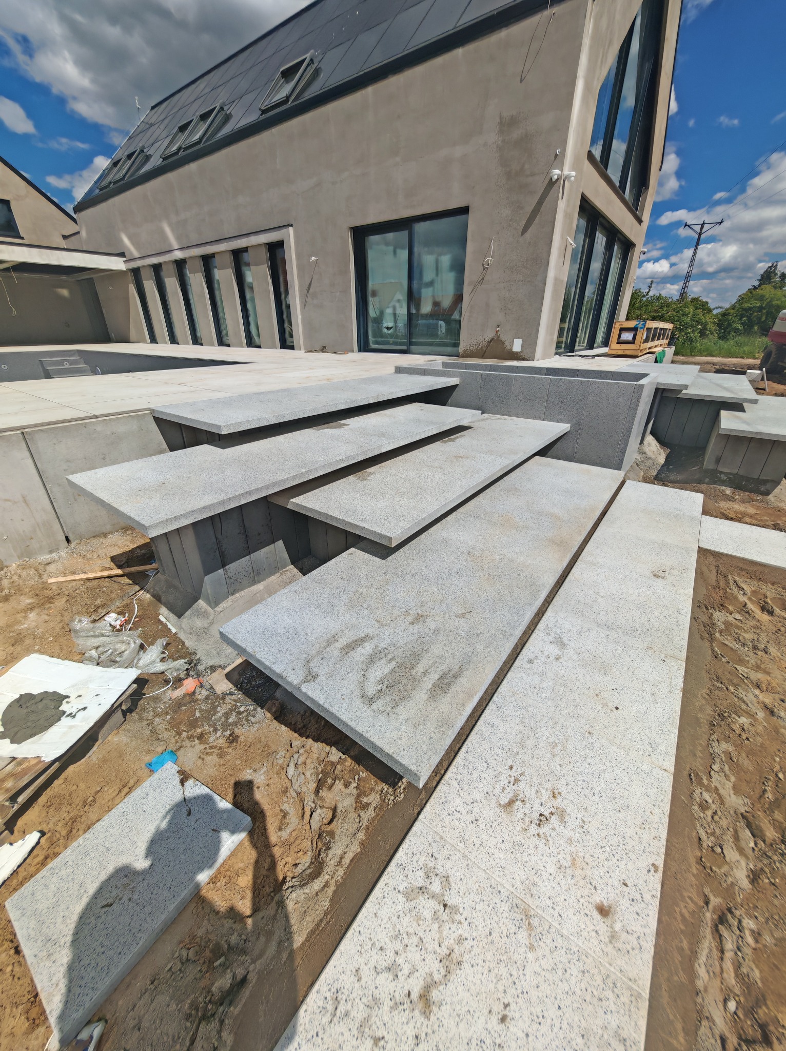 Granitowe schody zewnętrzne prowadzące do nowoczesnego domu z dużymi oknami. Widoczne płyty granitowe i elementy konstrukcyjne w trakcie montażu na zewnątrz budynku.