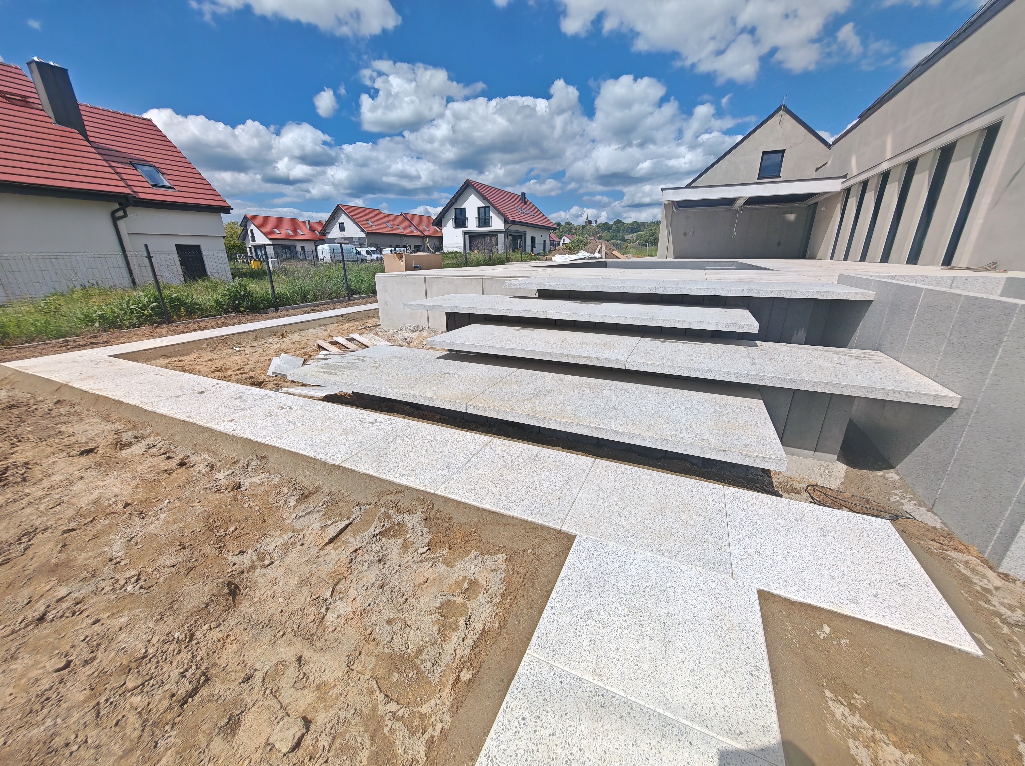 Nowoczesne schody z jasnej kostki granitowej prowadzą do domu na tle błękitnego nieba z chmurami. Widoczny fragment tarasu w trakcie budowy i domy w oddali.