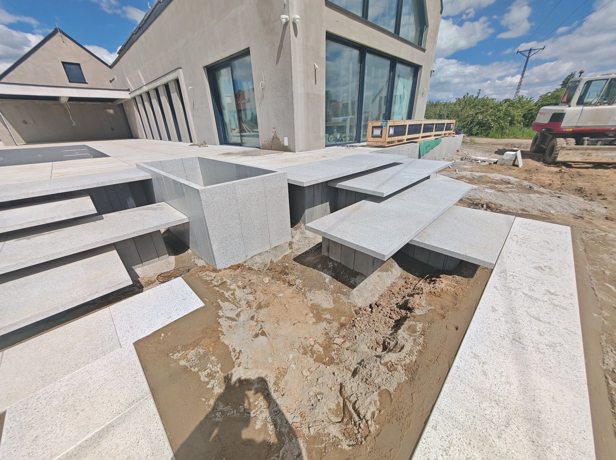 Nowoczesne schody i taras z jasnej kostki granitowej, w trakcie montażu przy domu z dużymi oknami. Widoczny sprzęt budowlany w tle.