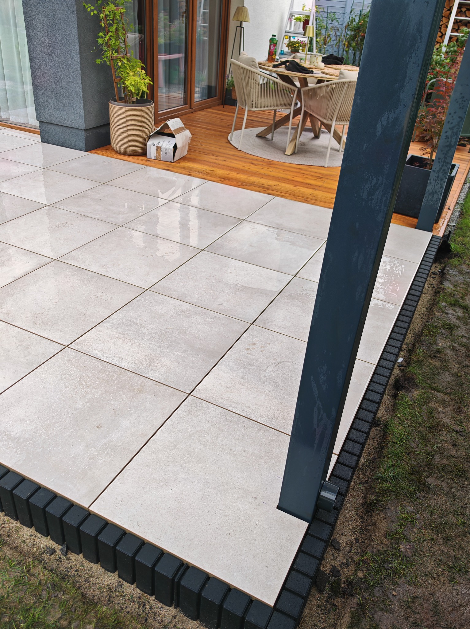 Jasne, kwadratowe płytki tarasowe z ciemną obwódką, fragment drewnianego tarasu z meblami ogrodowymi i pergolą w tle. Widok na świeżym powietrzu po deszczu.