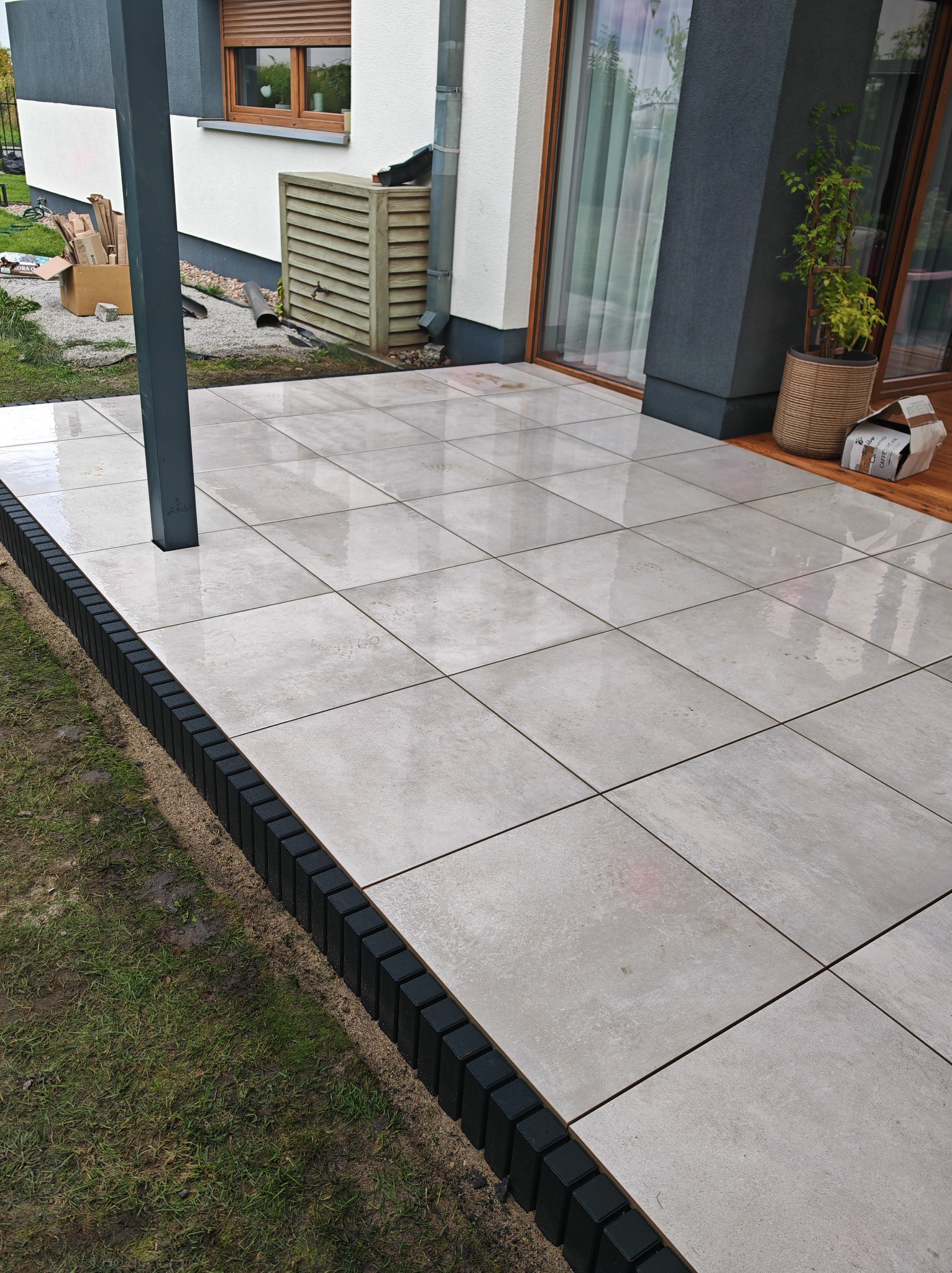 Ułożona, beżowa nawierzchnia z płytek na tarasie domu z czarnym obrzeżem. Widoczny fragment metalowej konstrukcji zadaszenia i trawnik.