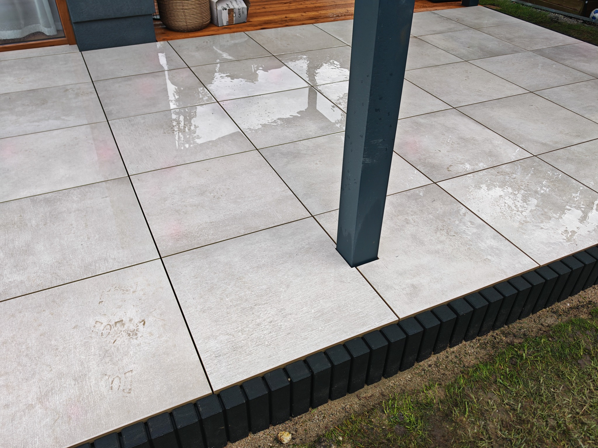 Szare, kwadratowe płyty tarasowe ułożone na zewnątrz, obrzeża z ciemnych palików, szara kolumna. Widoczne ślady po deszczu.