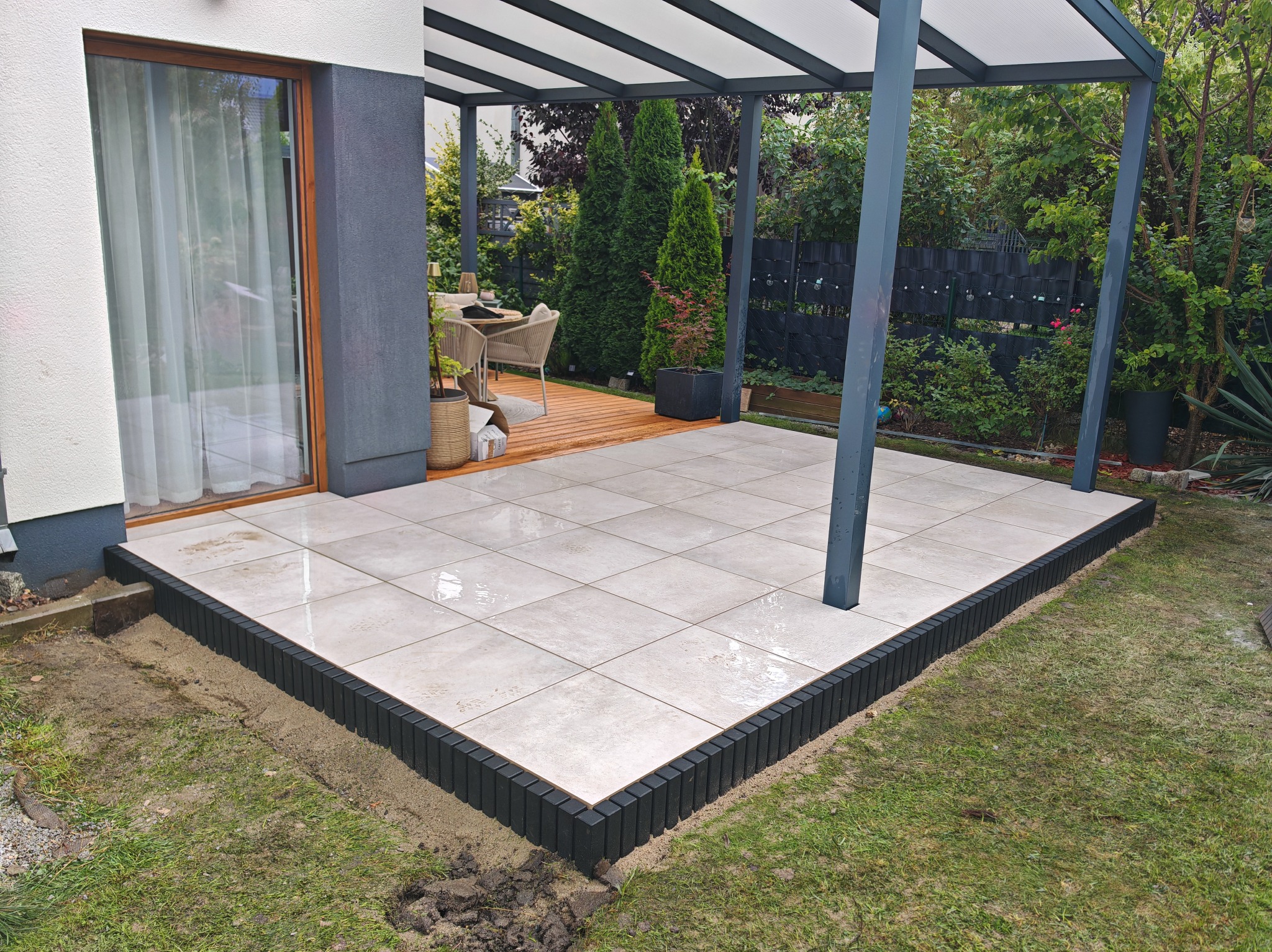 Nowoczesny taras z jasnych, kwadratowych płyt, obramowany czarnym, palisadowym obrzeżem. Zadaszenie pergoli, drewniany podest w tle i zielony trawnik.