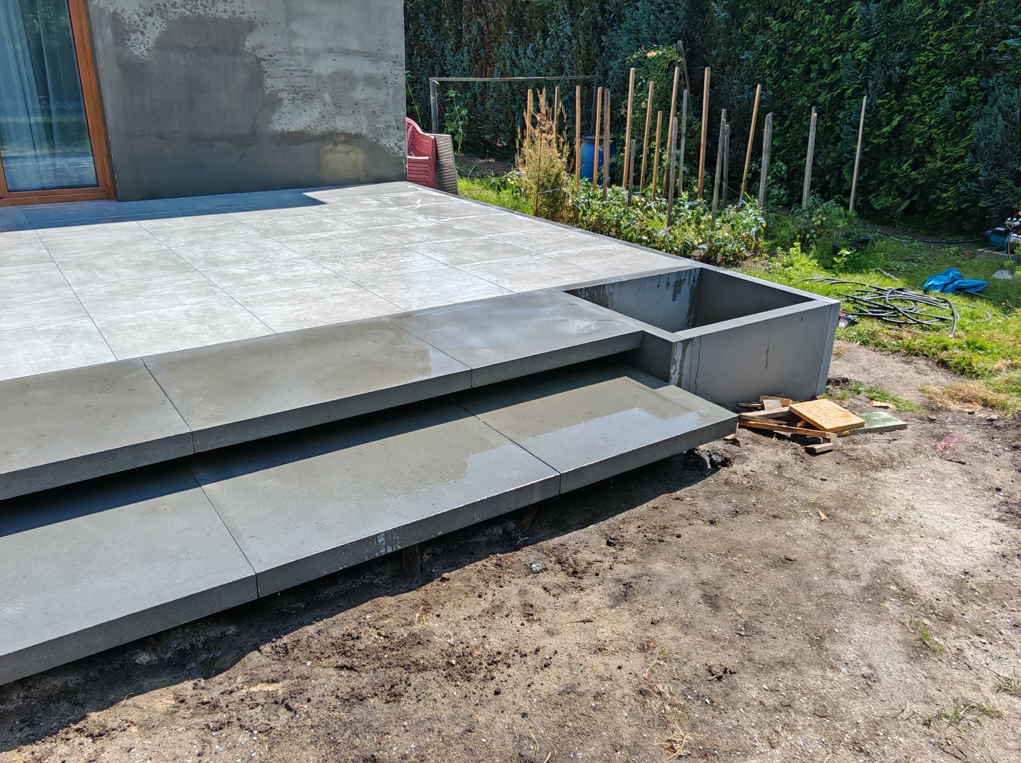 Nowoczesny taras z szarych płyt, dwustopniowe schody z betonu, obok rabata. Widoczne narzędzia i materiały budowlane. Tło: zieleń ogrodu, ściana budynku.