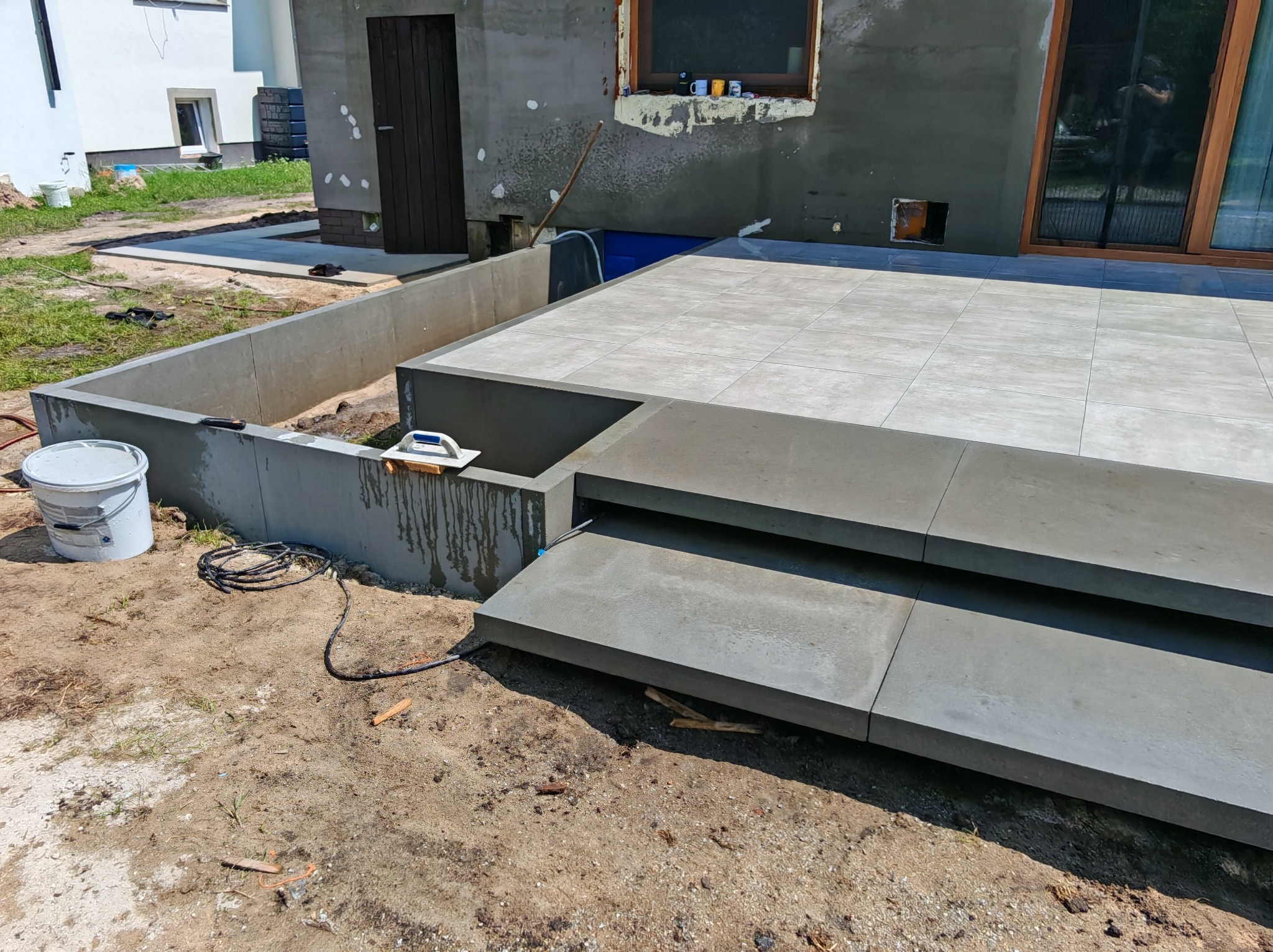 Wykończony taras z szarych płyt ceramicznych, z dwoma stopniami i betonowym obramowaniem, widok na częściowo otynkowany budynek z oknami i drzwiami.